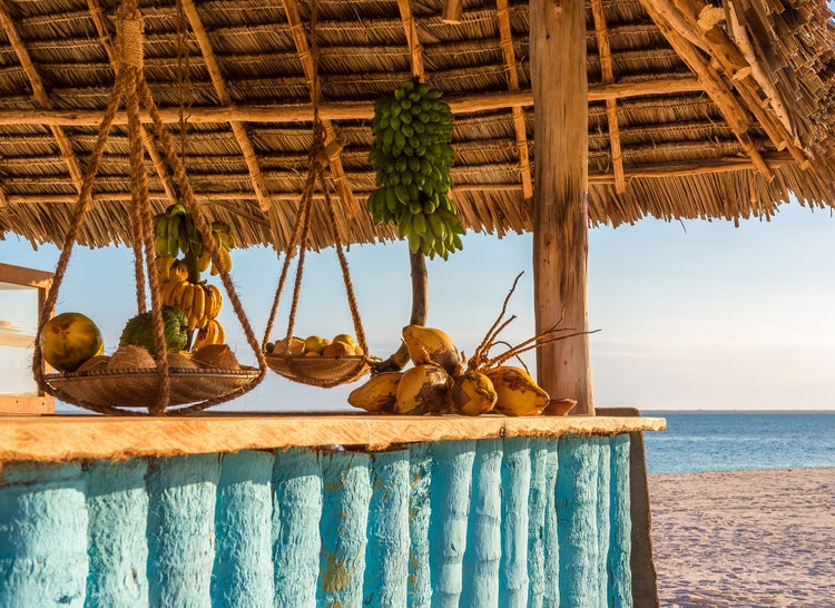 Außenbar mit Reetdach, Holztresen und hängenden Obstkörben aus Seil vor einer Strandkulisse.