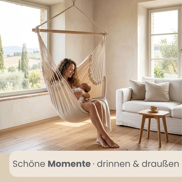 Frau mit Baby in einem hellen Hängesessel aus Stoff in einem lichtdurchfluteten Wohnzimmer mit Holzboden und Sofa.