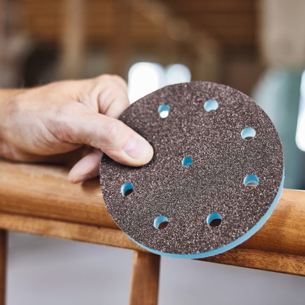 Hand hält ein rundes Schleifblatt mit Löchern vor einem Holzgeländer.