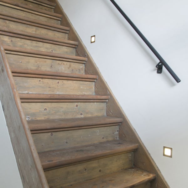 Holztreppe mit quadratischen Wandeinbauleuchten und einem schwarzen Metallhandlauf an einer weißen Wand.