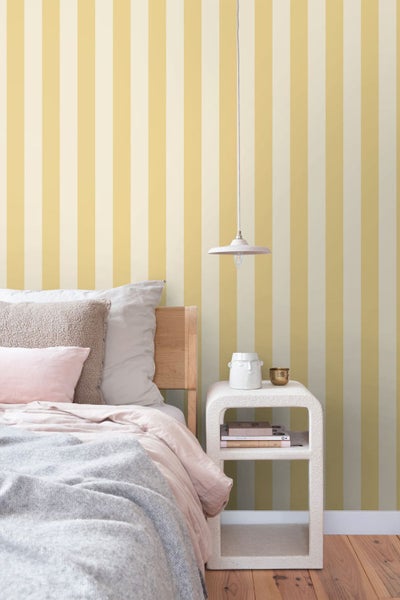 Helles Schlafzimmer mit vertikal gestreifter Tapete, einem Holzbett mit Kissen, einem weißen Nachttisch und einer Hängelampe.