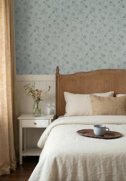 Schlafzimmer mit floral gemusterter Tapete und weißer Holzvertäfelung an der Wand. Ein Holzbett mit Rattankopfteil und ein weißer Nachttisch.