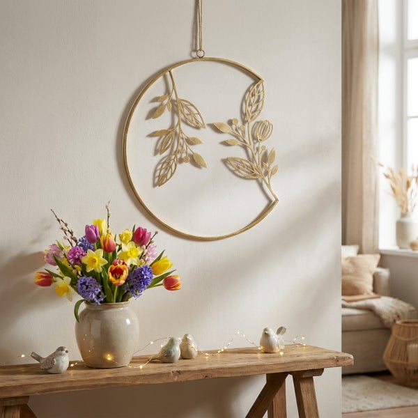 Wanddekoration aus Metall in Ringform mit Blattmotiv über einem Holztisch mit Frühlingsblumenstrauß und kleinen Dekovögeln.