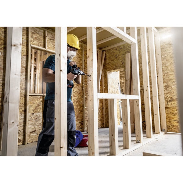 Mann mit Schutzhelm und Atemschutzmaske bohrt mit einem Akkubohrer ein Holzgerüst