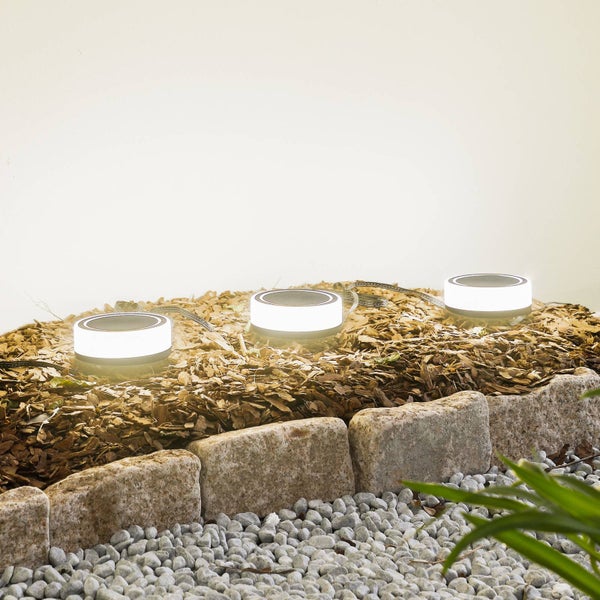 Drei runde LED-Gartenstrahler in einem Beet aus Rindenmulch, eingefasst von Natursteinen und hellem Kies.