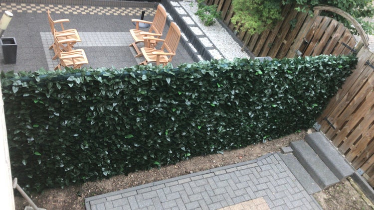 Sichtschutz aus künstlichem Efeu auf einer Terrasse mit Holzgartenstühlen und Steinboden.