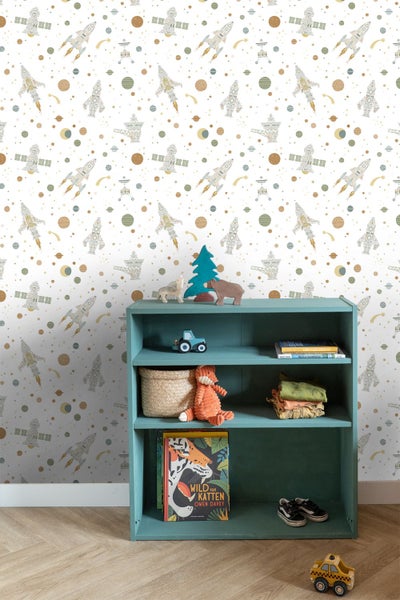 Vliestapete für Kinderzimmer mit Weltraum-Motiv aus Raketen und Planeten hinter einem Regal mit Spielzeug und Büchern in einem hellen Zimmer.
