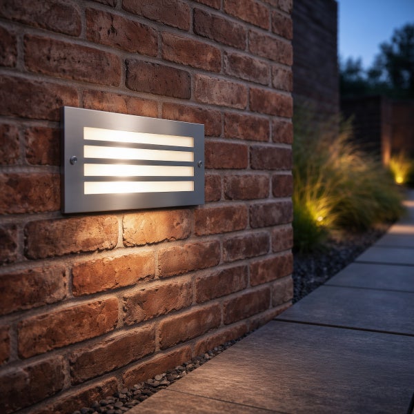 Rechteckige Einbauwandleuchte aus Metall mit horizontalen Lichtschlitzen an einer Ziegelwand über einem gepflasterten Gehweg.