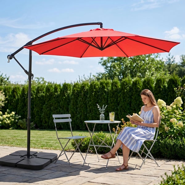Großer roter Ampelschirm in einem hellen Garten über einem grauen Bistro-Set aus Metall. Eine Frau liest auf einer Terrasse unter blauem Himmel.