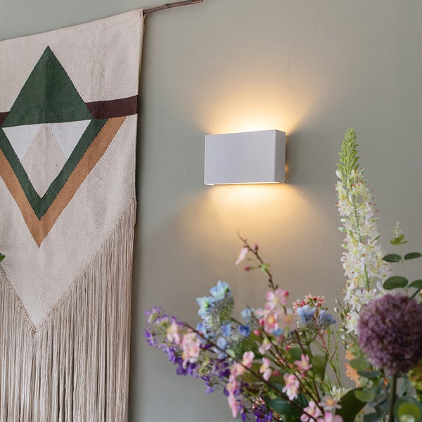 Rechteckige Wandleuchte neben Wandteppich und Blumenstrauß