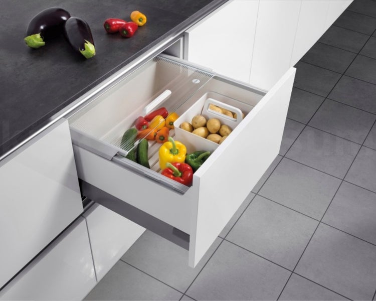Offene Küchenschublade mit Gemüseeinsatz und durchsichtigem Deckel, befüllt mit Paprika und Kartoffeln in einer modernen weißen Küche.