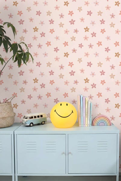 Tapete mit Blumenmuster vor einem Kinderzimmer mit Metallschrank, Smiley Lampe, Spielzeugbus, Büchern und Regenbogen Deko.