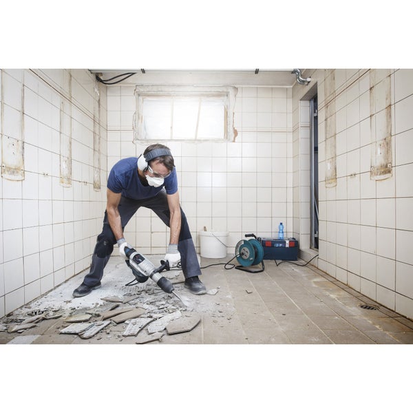 Ein Mann mit Schutzbrille, Gehörschutz und Staubmaske arbeitet mit einem Abbruchhammer in einem Raum mit Fliesen.