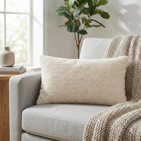 Rechteckiges Dekokissen mit weichem Kunstfell auf hellgrauem Sessel, mit grober Strickdecke und Beistelltisch in hellem, modernem Ambiente.
