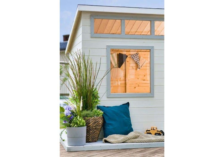 Modernes Gartenhaus aus weißem Holz mit grauen Fensterrahmen, Pflanzen, blauem Kissen und Spielzeug auf einer Holzterrasse.