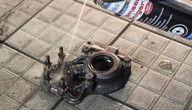 Reinigung eines verschmutzten Metallbauteils mit Bremsenreiniger aus einer Sprühdose mit E-COLL Logo auf einem Fliesenboden.
