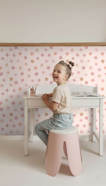 Ein glückliches Kind sitzt an einem Schreibtisch vor einer Kindertapete mit Blumenmuster in einem hellen, freundlich eingerichteten Kinderzimmer.