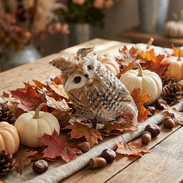 Herbstliche Tischdekoration mit einer Eulenfigur, kleinen Zierkürbissen, Ahornblättern, Kiefernzapfen, Kastanien und Eicheln auf Holzuntergrund.