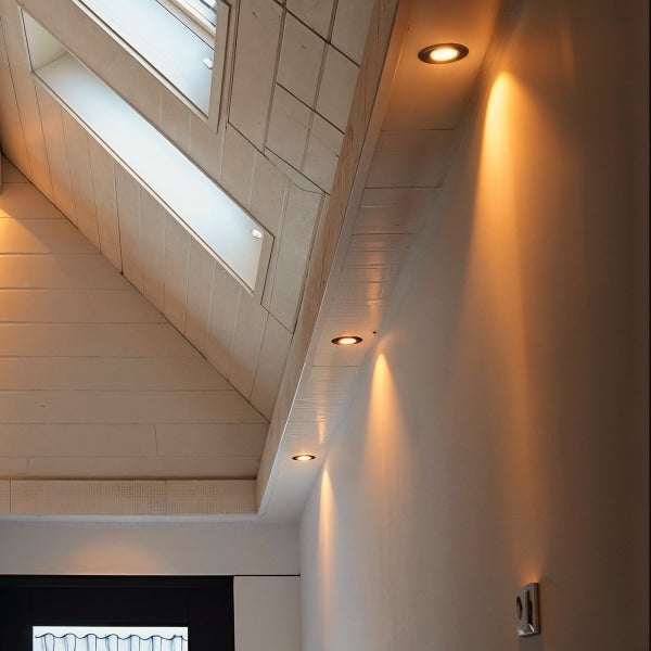 Modernes Zimmer mit Dachschräge, hellen Holzpaneelen an der Decke und eingebauten LED-Strahlern, die warmes Licht an die Wand werfen.