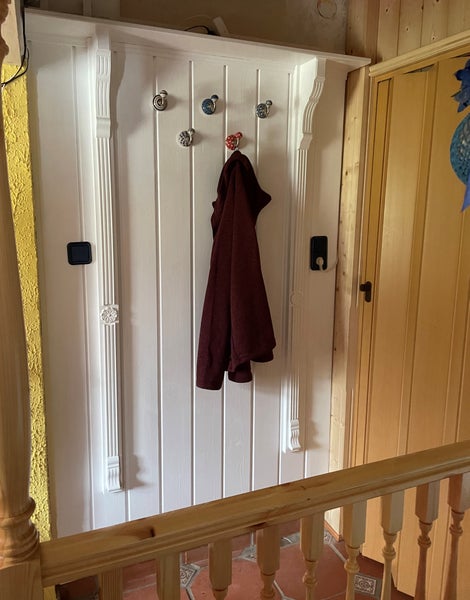 Weiße Wandgarderobe mit dekorativen Elementen und mehreren Kleiderhaken, an einem Haken hängt eine dunkle Jacke, im Flurbereich mit Holzgeländer.