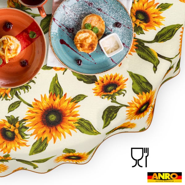 Tischdecke mit Sonnenblumen-Muster, dekoriert mit Tellern und Speisen. ANRO Deutsche Manufaktur Logo und Symbol für Lebensmittelechtheit.