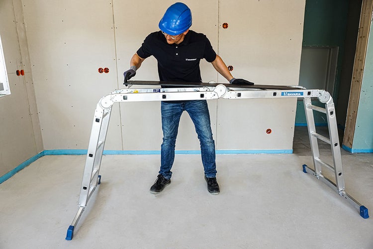 Ein Arbeiter mit Schutzhelm benutzt eine Arbeitsbühne in einem Raum mit Trockenbauwänden.