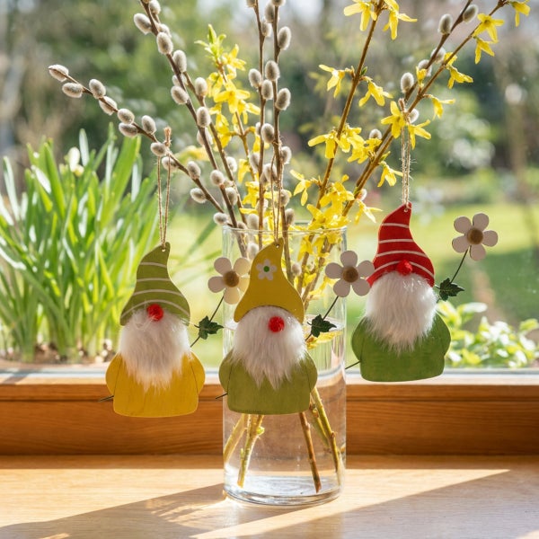Drei hängende Osterwichtel aus Holz mit Bart und Blumen an einer Glasvase mit Weidenkätzchen und Forsythienzweigen vor einem Fenster.
