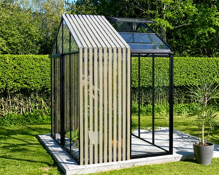 Modernes Gewächshaus aus Glas mit schwarzem Aluminiumrahmen und Holzlatten an einer Seite, im Garten auf einem Steinfundament stehend.