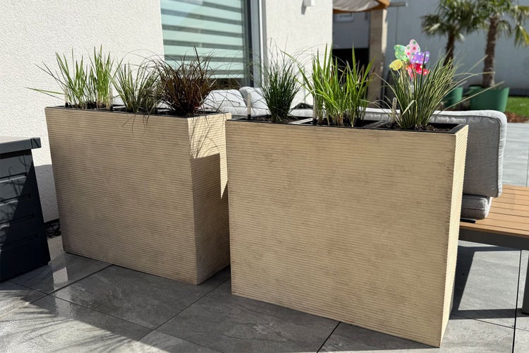 Zwei hohe rechteckige Pflanzkübel mit horizontaler Riffelstruktur auf einer Terrasse, bepflanzt mit verschiedenen Ziergräsern.