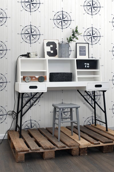 Weißer Schreibtisch mit Ablagefächern und grauem Hocker auf Holzpalette vor Wand mit Kompassmuster