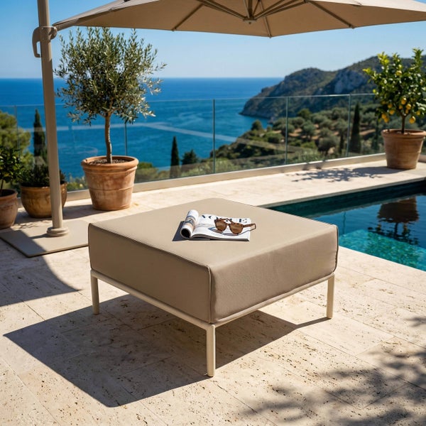 Quadratischer Gartenhocker mit beigem Polster auf einer hellen Steinterrasse am Pool mit Meerblick und Sonnenschirm.