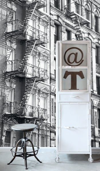 Szene mit einem Hocker, einem Rollschrank mit '@' Symbol und einem 'T' Symbol vor einem Gebäude.