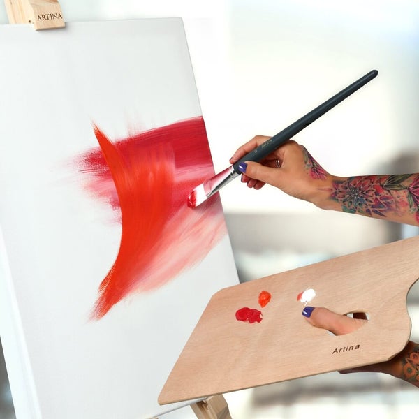 Eine Person malt mit einem Pinsel und Farbe auf eine Leinwand, wobei eine Holzpalette zur Mischung der Farben verwendet wird.