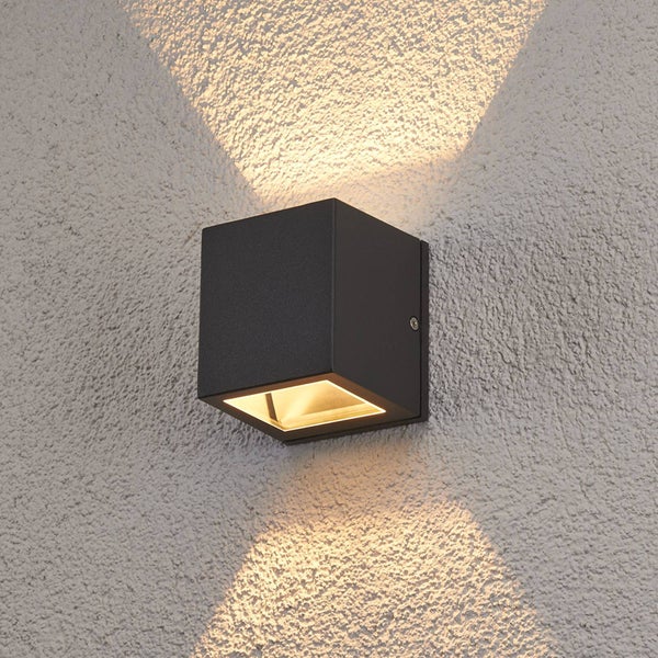 Würfelförmige Außenwandleuchte in Schwarz mit Lichtkegel nach oben und unten an einer strukturierten Wand.