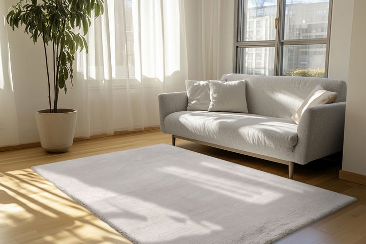 Helles, modern eingerichtetes Wohnzimmer mit grauem Sofa, flauschigem Teppich, Holzboden und großer Zimmerpflanze in einem hellen Raum.