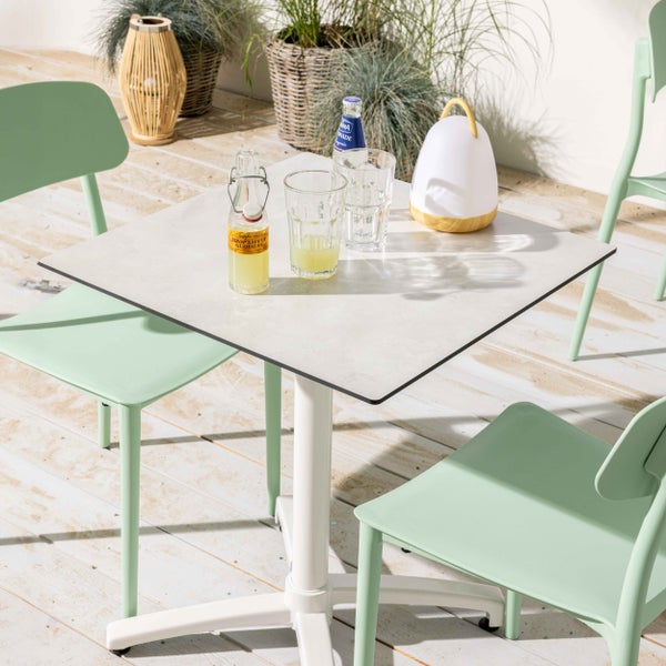 Quadratischer Bistrotisch in Steinoptik mit weißem Standfuß und mintgrünen Gartenstühlen auf einer Holzterrasse.