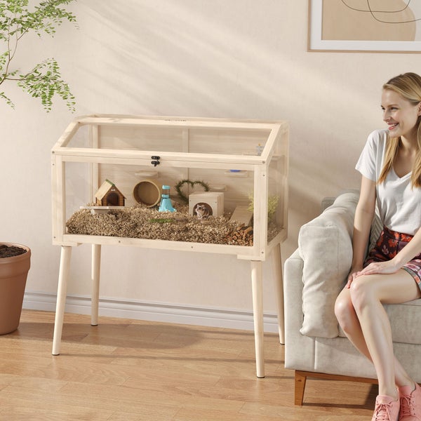Modernes Hamstergehege aus hellem Holz auf Beinen, eingerichtet mit Streu und Zubehör in einem hellen Wohnzimmer.