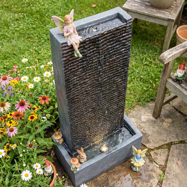 Gartenbrunnen in Steinoptik mit Wasserwand, Feenfiguren und Hasenfigur in einem Garten mit Blumen.