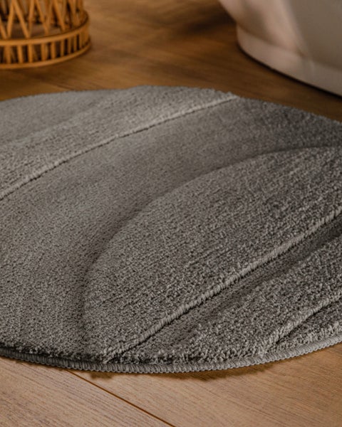 Runder, strukturierter Badeteppich mit geprägten Linien auf einem Holzboden neben einer Badewanne und einem Korbgeflecht.