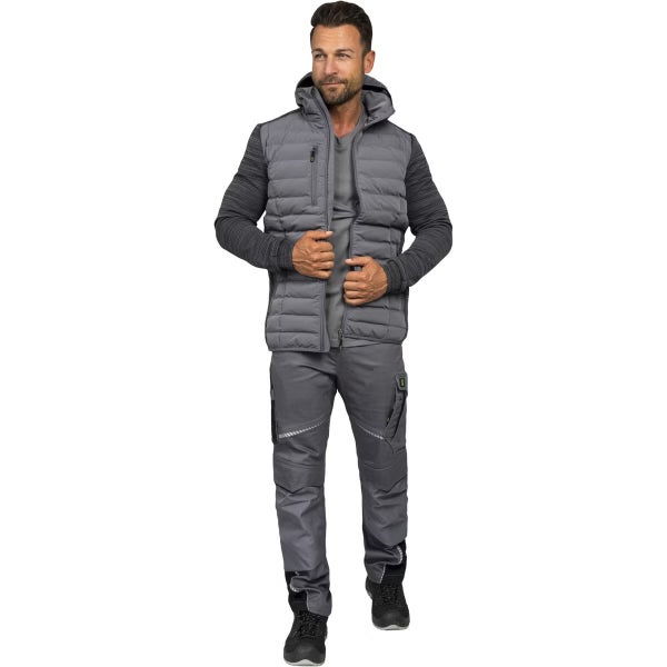 Mann in grauer Arbeitskleidung bestehend aus einer gesteppten Weste mit Kapuze, einem Langarmshirt, einer Arbeitshose und schwarzen Arbeitsschuhen.