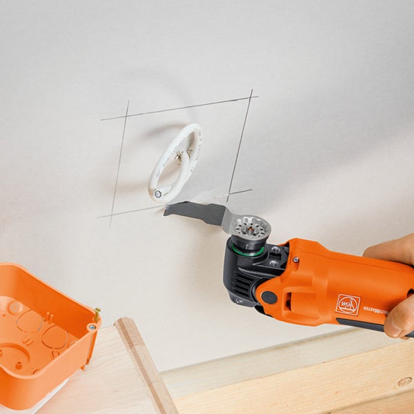 Oszillierendes Elektrowerkzeug schneidet eine Öffnung in eine Gipskartonwand in der Nähe einer elektrischen Leitung, um eine orangefarbene Anschlussdose freizulegen.