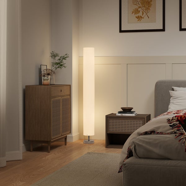 Modernes Schlafzimmer mit warm leuchtender, zylindrischer Stehleuchte sowie Kommode und Nachttisch aus Holz mit Rattangeflecht.