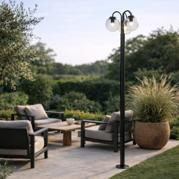 Dreiflammige Außenstehleuchte aus schwarzem Metall mit gläsernen Kugeln in einem hellen, modern gestalteten Garten mit Sitzgruppe.