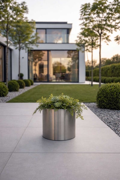 Zylindrisches Pflanzgefäß aus Edelstahl mit grünen Pflanzen auf einer modernen Terrasse mit gepflegtem Garten und Haus im Hintergrund.