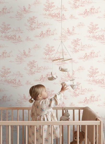 Kleinkind in Holzgitterbett vor Wand mit Toile-de-Jouy-Tapete mit ländlichen Motiven, helles Kinderzimmer mit hängendem Mobile.