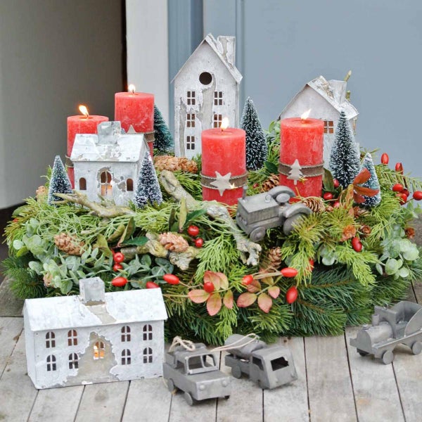 Adventskranz mit vier roten Stumpenkerzen, kleinen Zinkhäusern, Miniatur-Tannenbäumen und grauen Spielzeugautos auf Tannenzweigen.