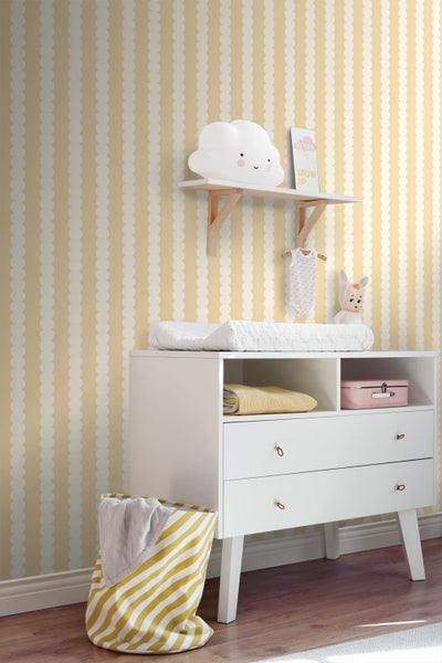 Kinderzimmer mit gelb-weiß gemusterter Vliestapete, weißer Wickelkommode und Wandregal.