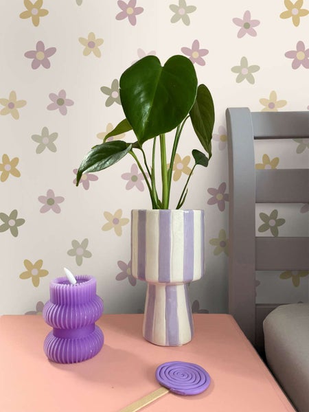 Eine Pflanze in einem gestreiften Übertopf steht auf einem Tisch vor einer Blumentapete neben einer gerippten Kerze und einem runden Dekoelement.
