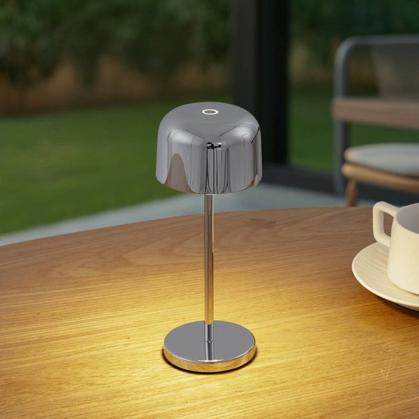 Moderne kabellose Tischleuchte aus verchromtem Metall mit warmem Licht auf einem Holztisch im Außenbereich.