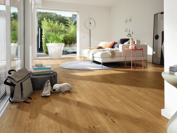 Helles Wohnzimmer mit Holzoptik Bodenbelag, einem Sofa, einem runden Teppich und einem großen Fenster mit Blick auf eine Terrasse.